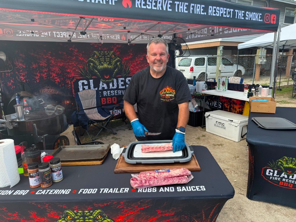Pitmaster trimming a rack of ribs at the catering tent, under a banner reading competition BBQ, catering, food trailer, rubs and sauces