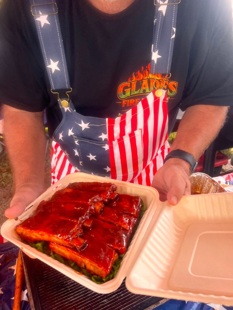 Pitmaster in a stars and stripes apron holding an open box of glazed ribs