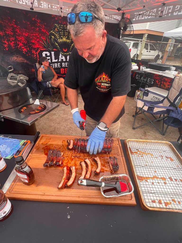 Pitmaster in gloves slicing a glazed rack of ribs on a wood board at the competition tent