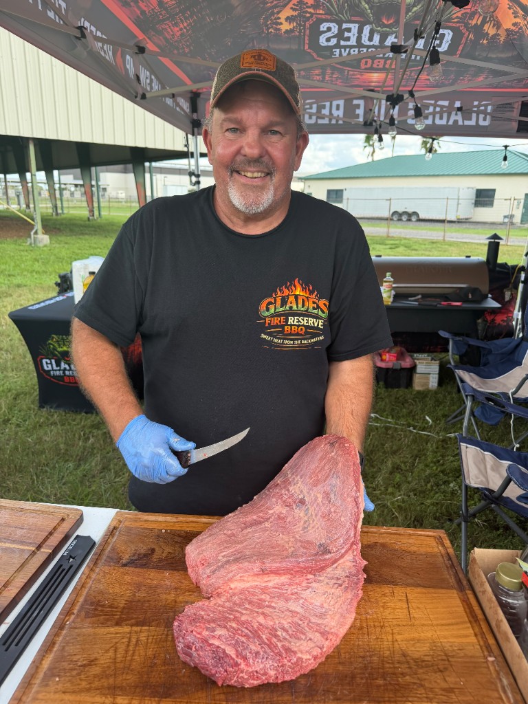 Smiling pitmaster with a boning knife behind a whole raw packer brisket on a wood board