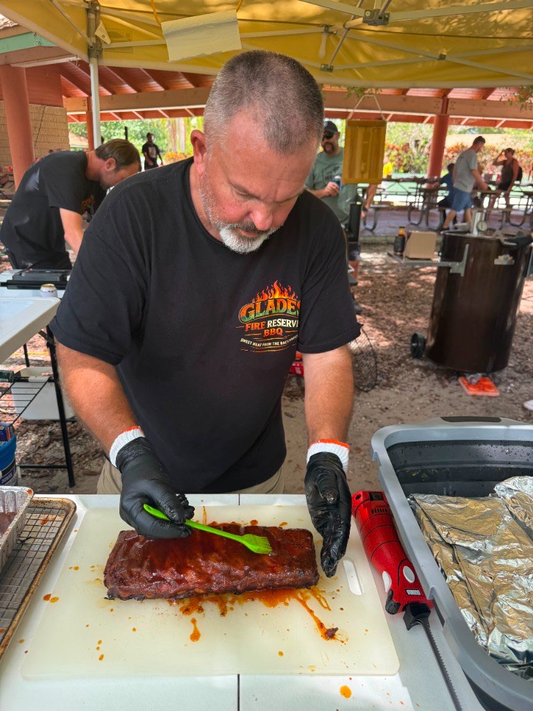 Pitmaster brushing a final coat of glaze onto a rack of ribs under a park pavilion