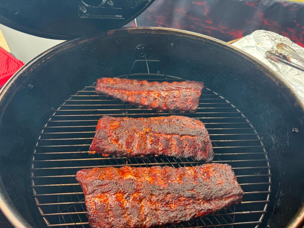 Three racks of ribs with a dark bark resting on the grate of a kettle cooker