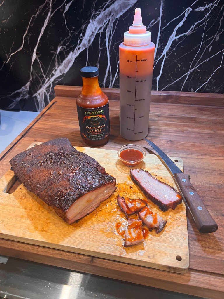 Sliced smoked pork belly on a board next to a bottle of Glades Competition Glaze