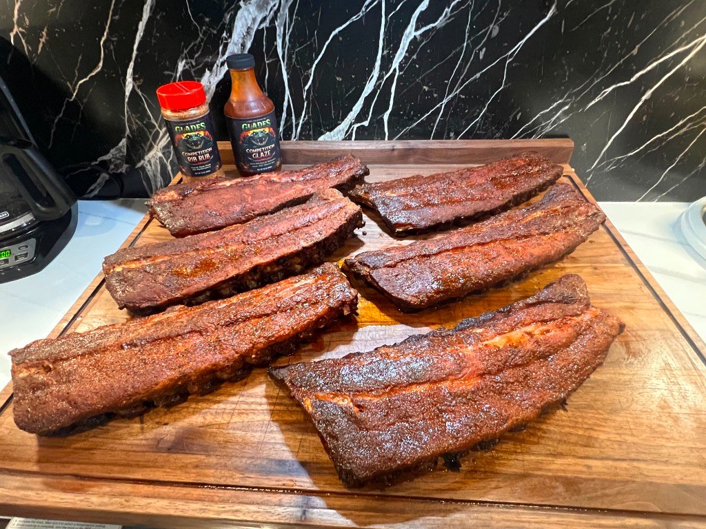 Six smoked racks of ribs spread across a walnut board behind bottles of Glades Competition Rib Rub and Glaze