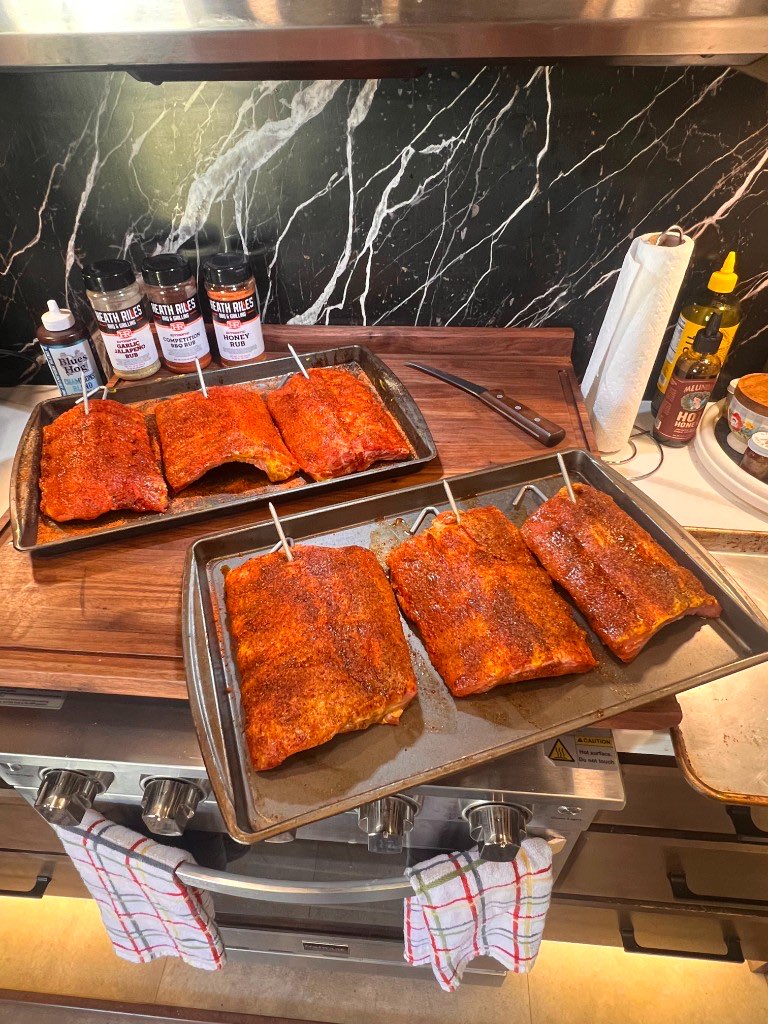 Two sheet pans of ribs coated in rub on a kitchen counter, hangers already set in the racks