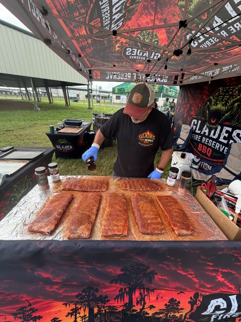 Pitmaster shaking seasoning over eight racks of ribs laid out on foil under the Glades Fire Reserve canopy