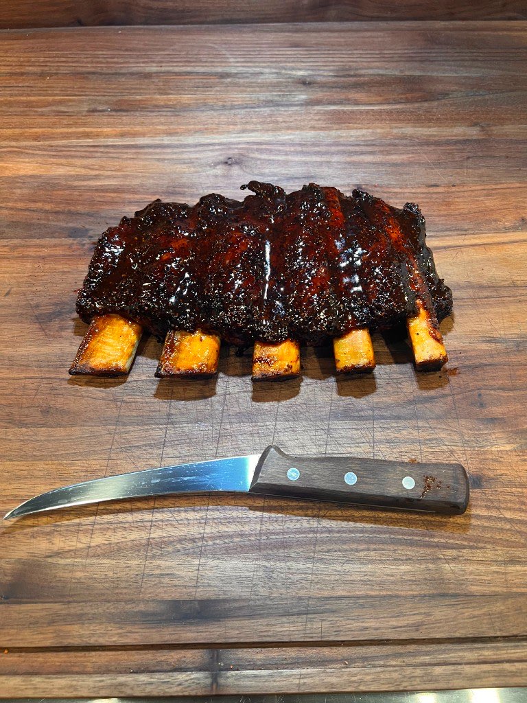 A finished rack of ribs sliced into clean bones on a walnut board beside a boning knife
