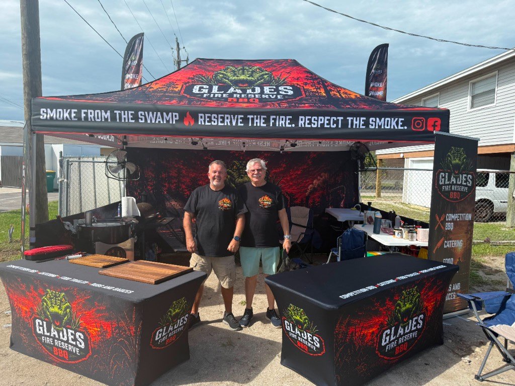 Two team members in front of the Glades Fire Reserve canopy and skirted tables, printed with Smoke from the Swamp — Reserve the Fire. Respect the Smoke.