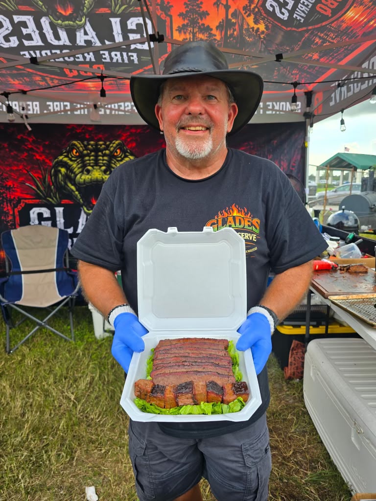 Pitmaster in a black leather hat holding up an open turn-in box of sliced brisket and burnt ends at the Glades Fire Reserve tent